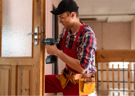 Carpenter in red overalls using a drill to repair a wooden door indoors.