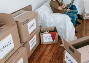 Cardboard boxes labeled Office, Winter Clothes, and Summer Clothes in a room with wooden flooring and a seated person.