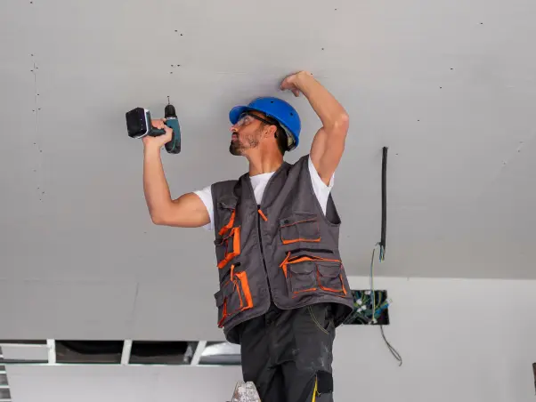 Construction worker in hard hat and vest using a drill on ceiling indoors, performing installation.