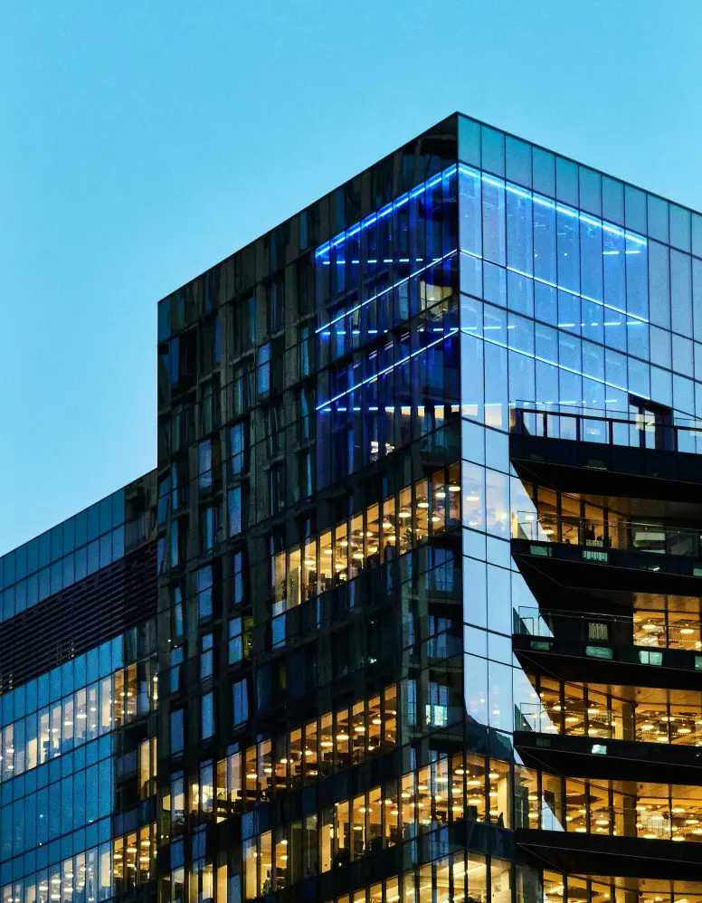 Modern glass skyscraper with glowing blue lights and warm office interiors at dusk against a clear sky.