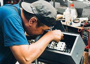 Technician repairing electronic device in workshop, wearing a cap and blue shirt, focused on fixing internal components.