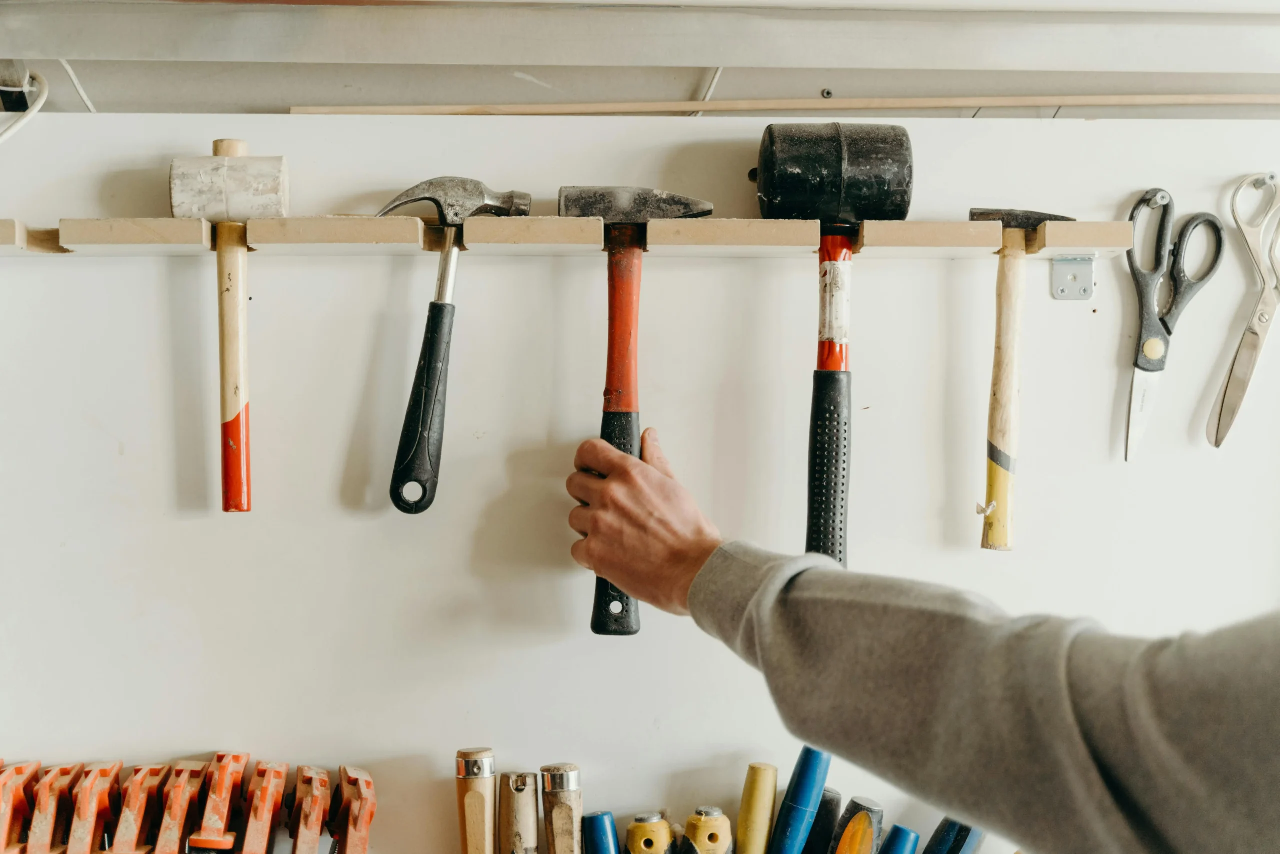 Hand reaching for a hammer on a workshop tool rack with various hammers and scissors hanging.