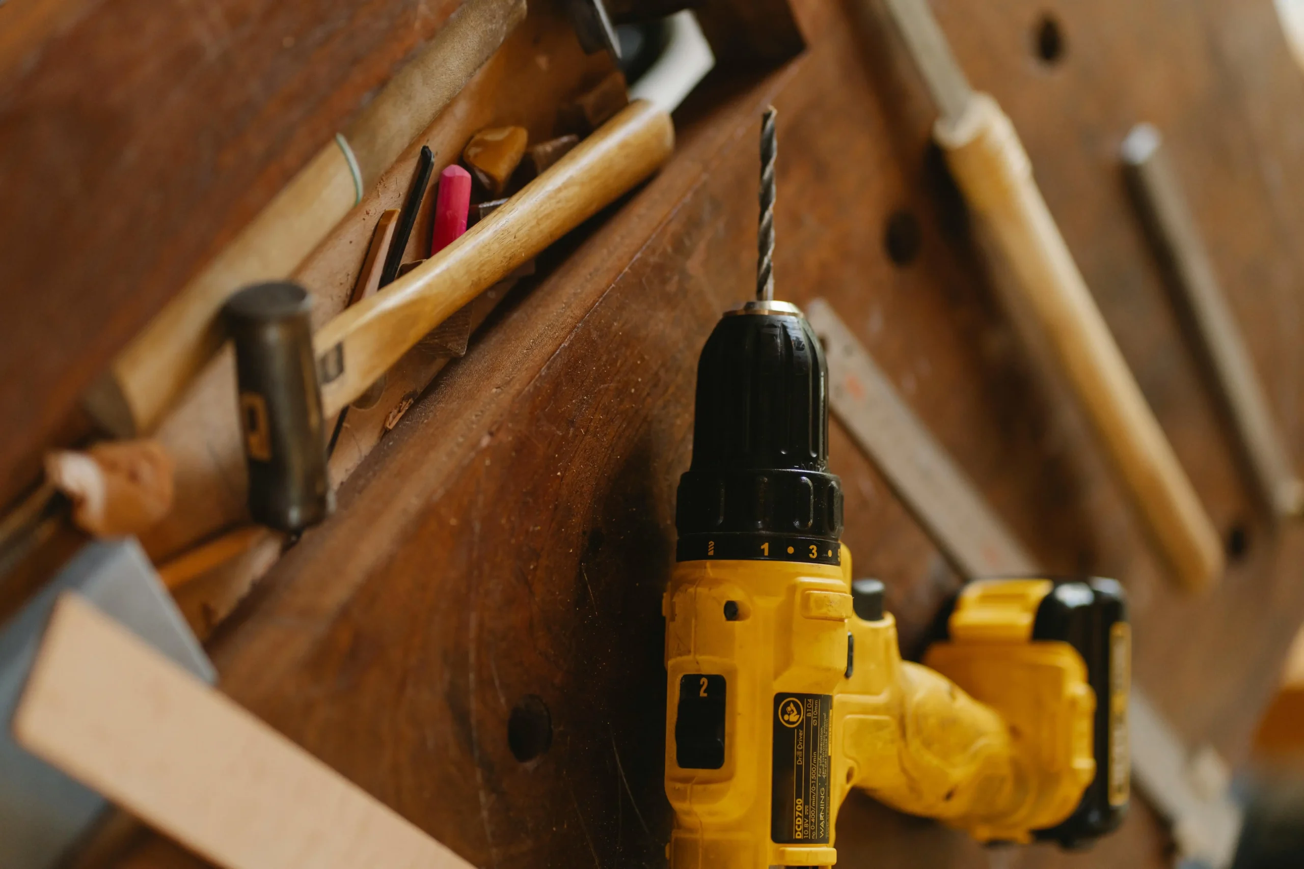 Yellow cordless drill and assorted hand tools on wooden workbench in a workshop setting.