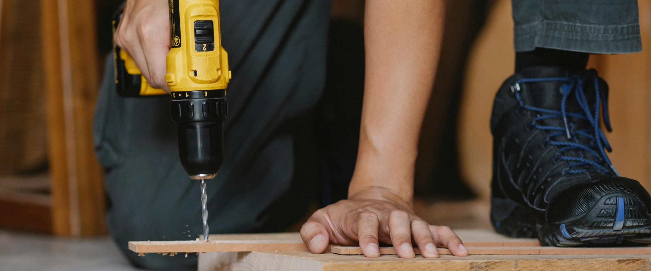 Person using a power drill on wood, wearing safety boots, focusing on DIY or construction project.