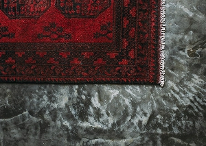 Close-up of a red and black patterned rug on a textured grey floor, showcasing intricate design and contrasting surfaces.