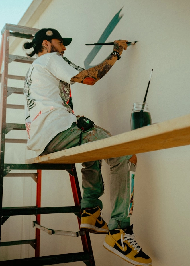 Tattooed artist on ladder paints mural with brush, wearing colorful sneakers and casual attire. Urban street art in progress.