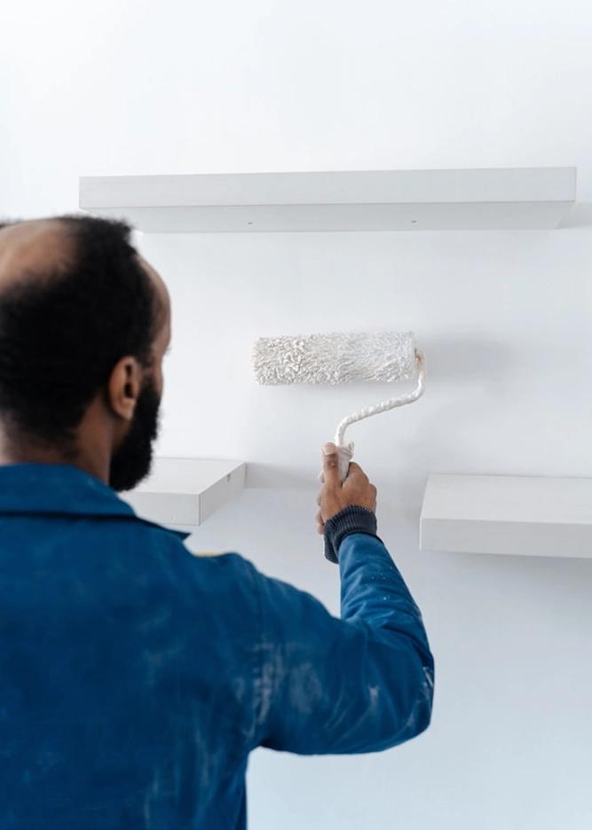 Man painting a white wall with a roller near shelves, wearing a blue jacket. Home renovation and interior decoration concept.