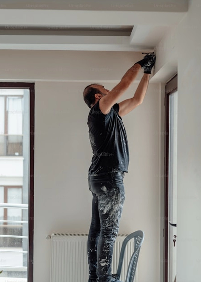 Person standing on chair to paint ceiling, wearing black clothes with paint stains in a bright room.