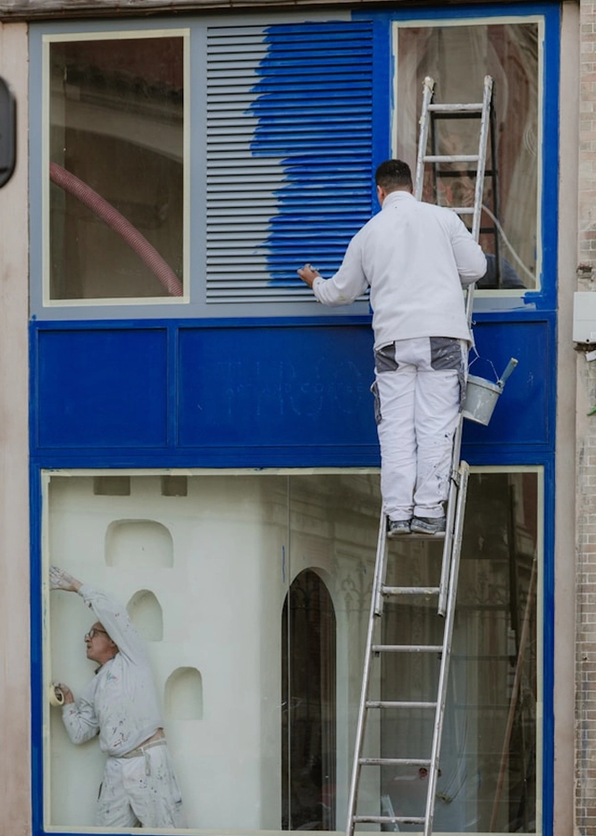 Painters renovating storefront with blue paint, one on ladder, another inside, creating vibrant exterior finish.