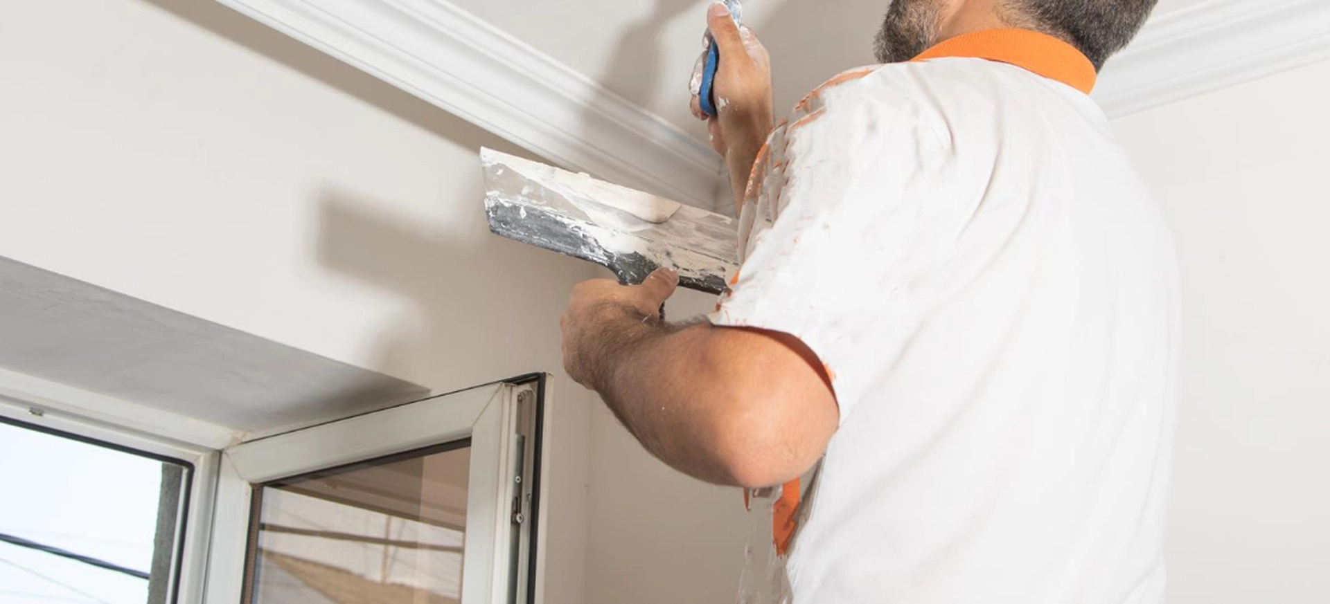 Worker applying plaster to ceiling corner near window for a smooth finish.