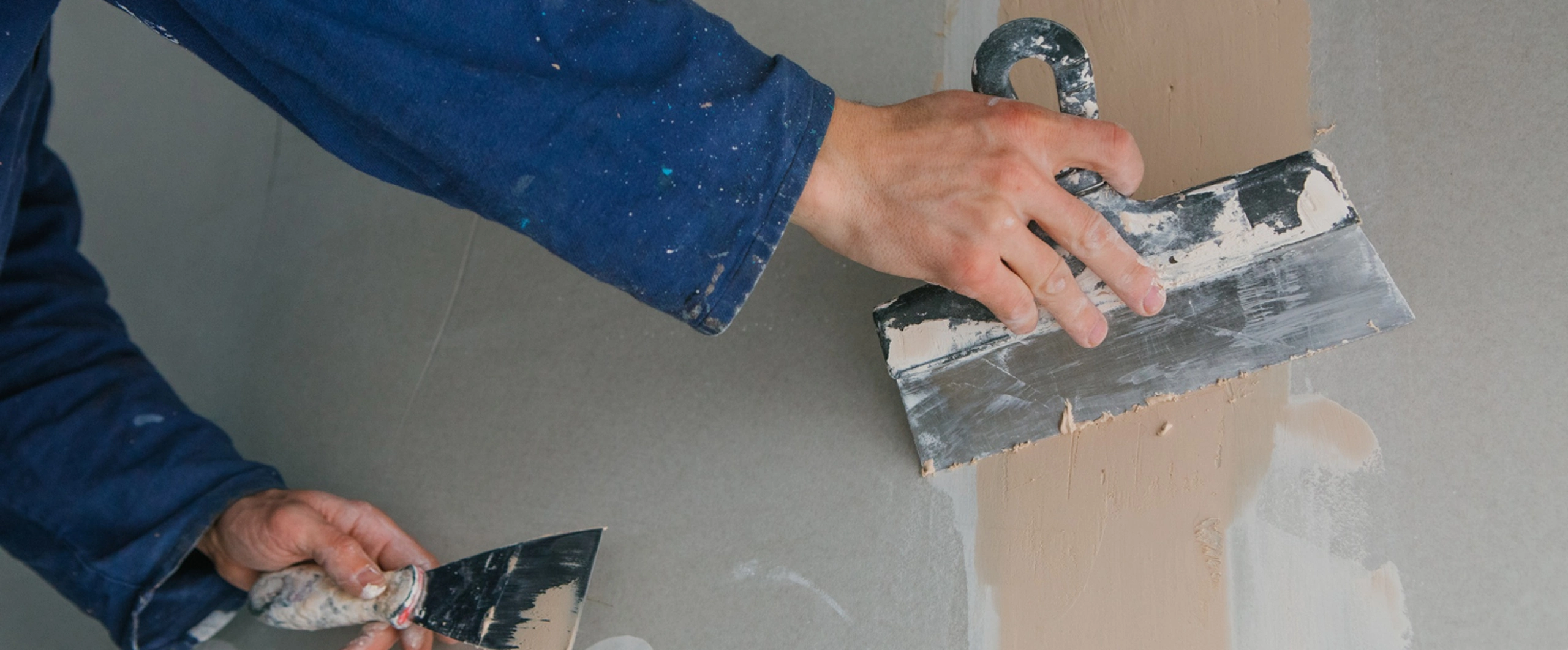 Person smoothing drywall with putty knives and joint compound, wearing blue sleeves.