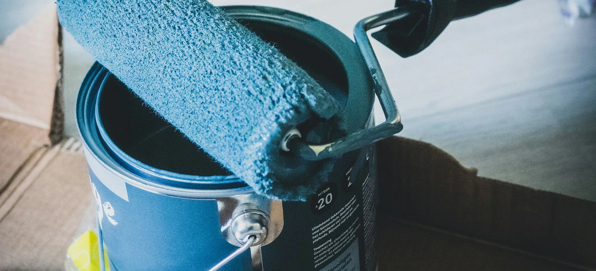 Paint roller coated with blue paint rests on open paint can inside a cardboard box.