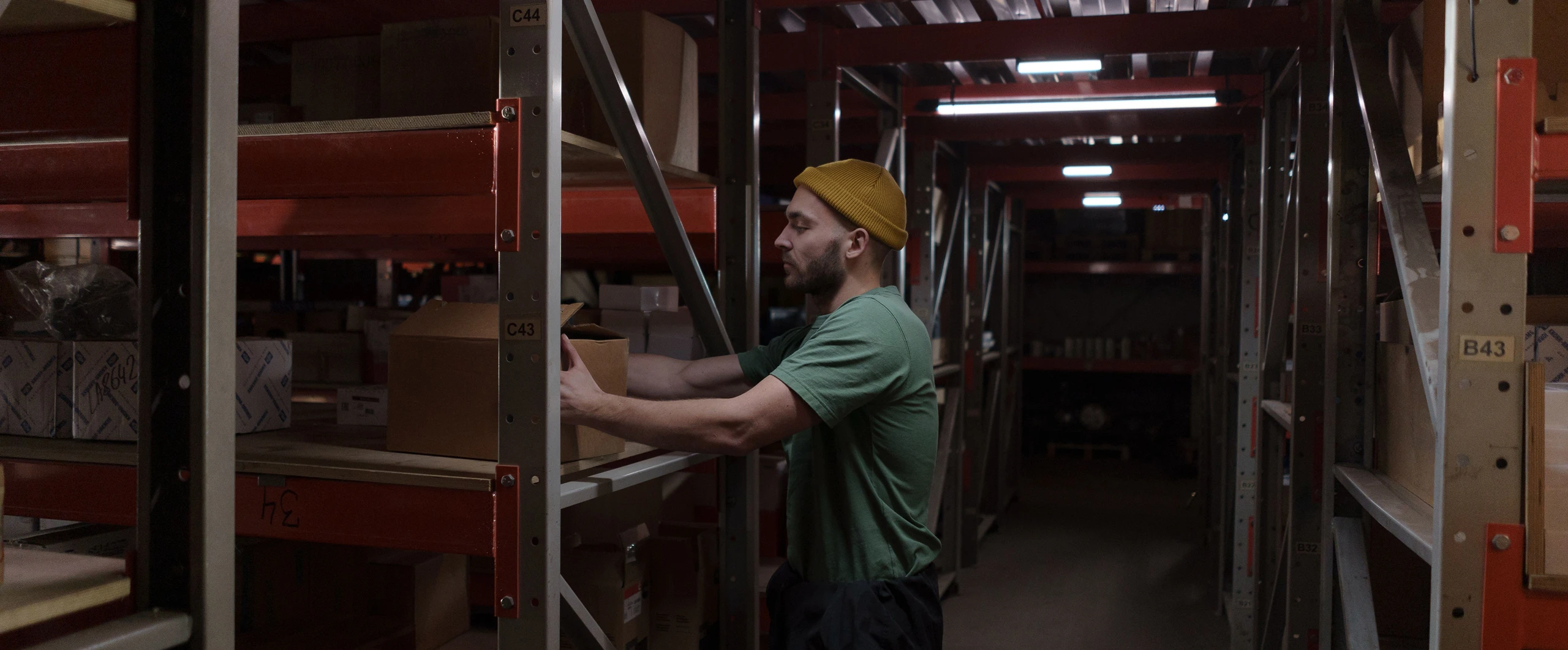 A man wearing a yellow hard hat and green shirt is arranging boxes on a shelf in a dimly lit warehouse.