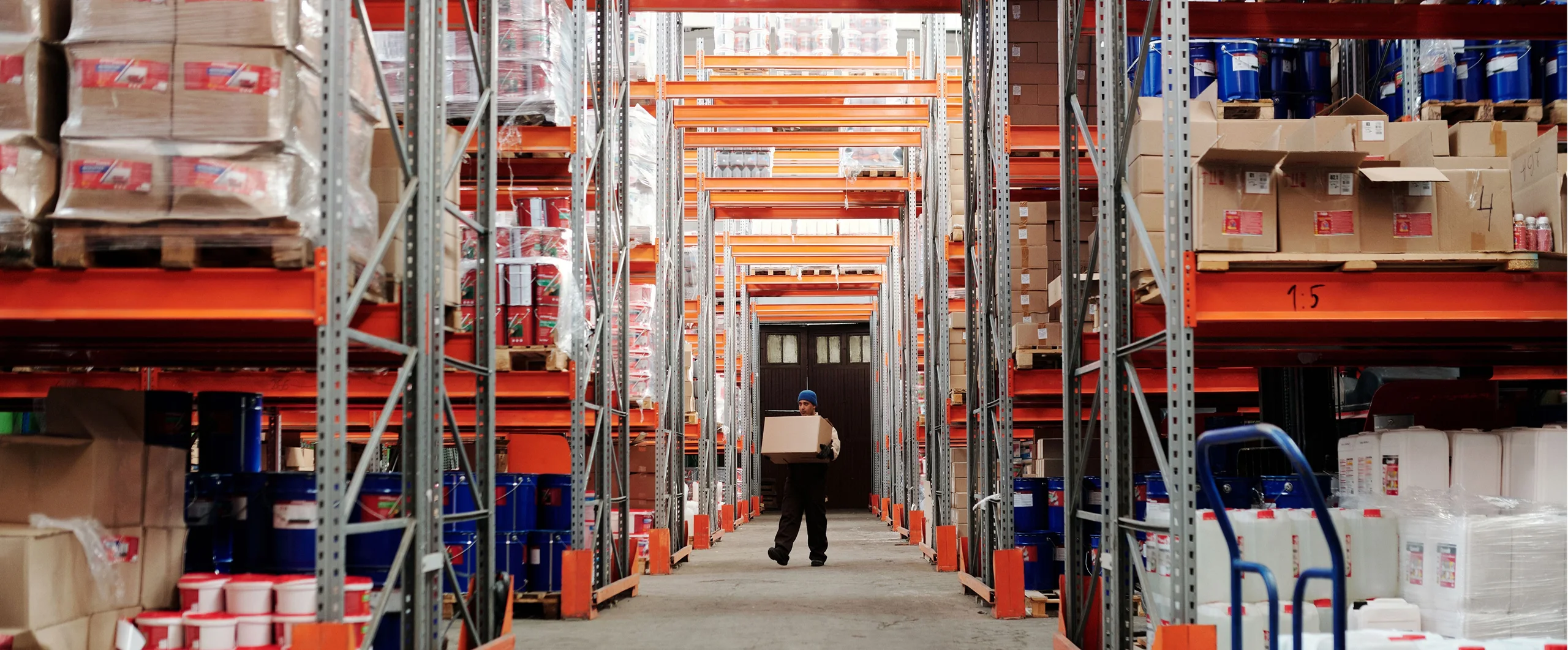A person carries a cardboard box down an aisle in a warehouse filled with shelves of various goods and containers.