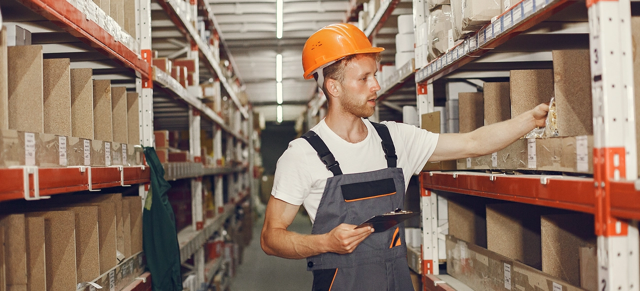 A worker in overalls and an orange helmet in a warehouse aisle checks inventory on shelves, holding a clipboard. The setting is organized and focused.