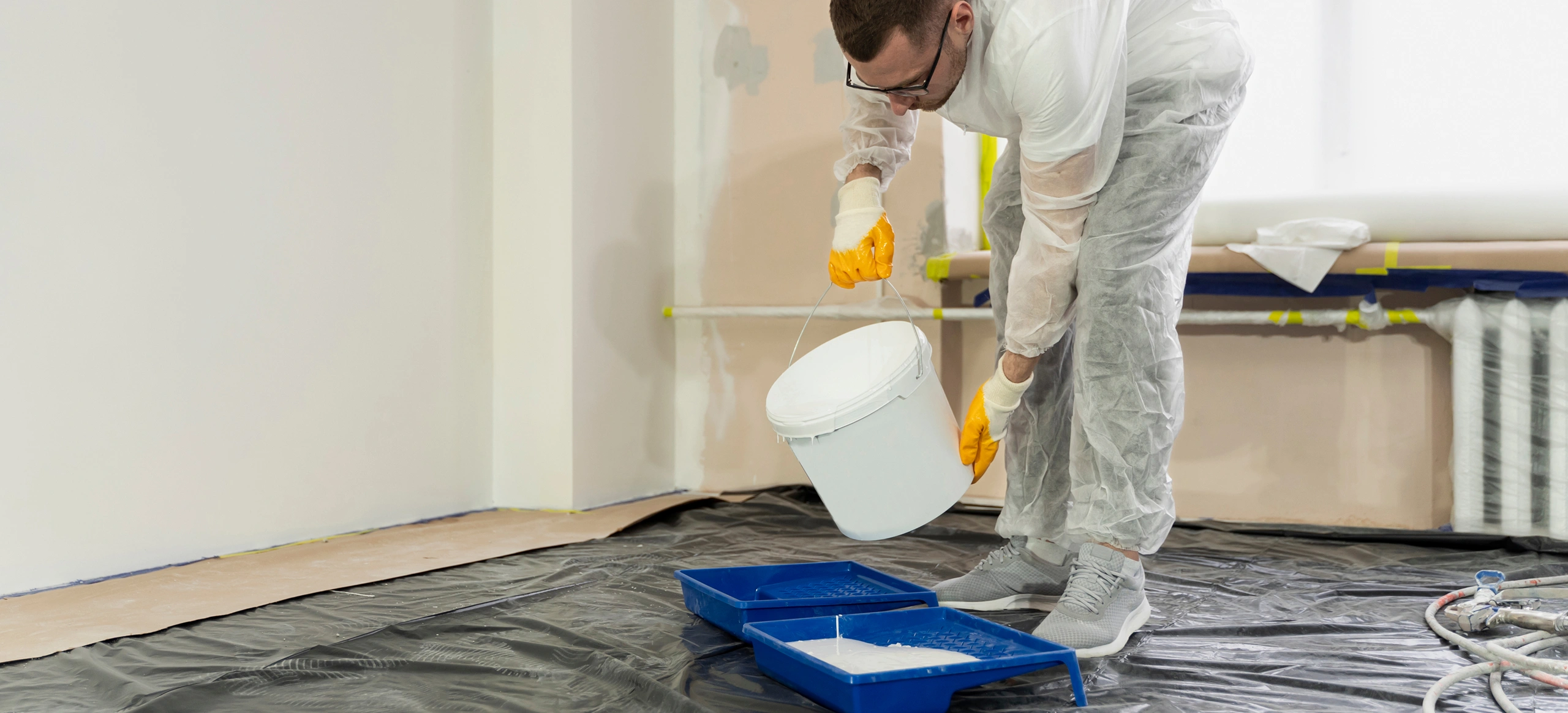 A person in protective gear pouring paint from a bucket into a blue paint tray on a covered floor during a room renovation.