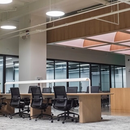 Modern office interior featuring a long wooden table with black ergonomic chairs, large windows, and bright overhead lighting.