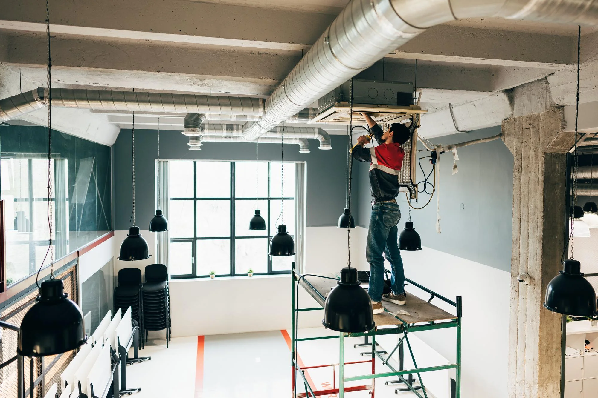 Technician on a scaffold tower servicing a ceiling-mounted air conditioning unit in an open-plan office