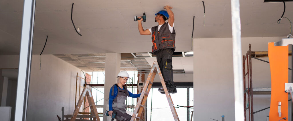 Two construction workers installing a drywall ceiling, one drilling from a ladder while a colleague steadies it below